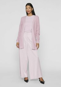 Cardigan rosa chiaro con lavorazione a maglia e maniche lunghe; camicia rosa chiaro abbinata e pantaloni a gamba larga; scarpe nere con tacco alto.