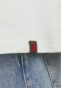 Hvid T-shirt med grøn label, der har en rød cirkel, præsenterer en glat tekstur og en syet kant, parret med denimstof.