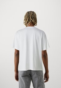 Valge puuvillane T-särk, millel on lõdva istuvuse, lühikesed varrukad ja sirge äärejoon. Omab minimaalset disaini, ilma silmapaistvate logode või mustriteta.