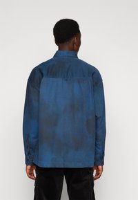 Blå denimjakke med en avslappet passform, med knappemansjetter og en subtil sky-lignende tekstur. Bakutsikten avslører distinkte fargevariasjoner.