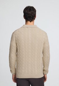 Beżowy sweter z długim rękawem typu cable-knit, z kołnierzem polo, prążkowanymi mankietami i wykończeniem u dołu. Teksturowany wzór z subtelnym połyskiem.