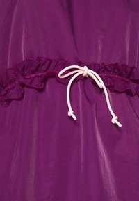 Lila tyg med en silkeslen yta, prydd med en rynkad remsa och en vit dragsko för justering i midjan.