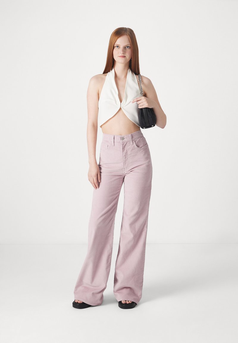 Haut court blanc à dos nu avec devant torsadé, pantalon large rose clair, et sac à main noir. Des sandales plates noires complètent le look.