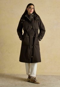 Hnedá parka s opaskom a vysokým golierom, s cvokmi a vreckami s klopami. Hladká textúra, dĺžka po kolená, vrstvená cez šedý top a biele nohavice.