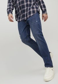 Jeans en denim bleu avec une coupe droite, comportant des détails déchirés et un lavage moyen. Associés à une chemise à carreaux et des baskets blanches.