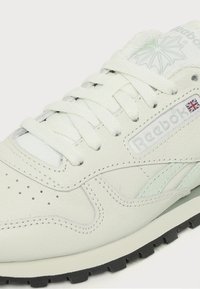 Gros plan d'une sneaker blanche en cuir Reebok avec lacets, logo de la marque, drapeau de l'Union Jack et semelle extérieure noire visible.