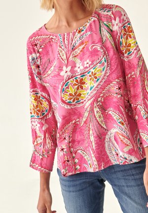 Femme portant un chemisier à manches longues rose vif avec des motifs floraux et cachemire colorés, associé à un jean bleu.