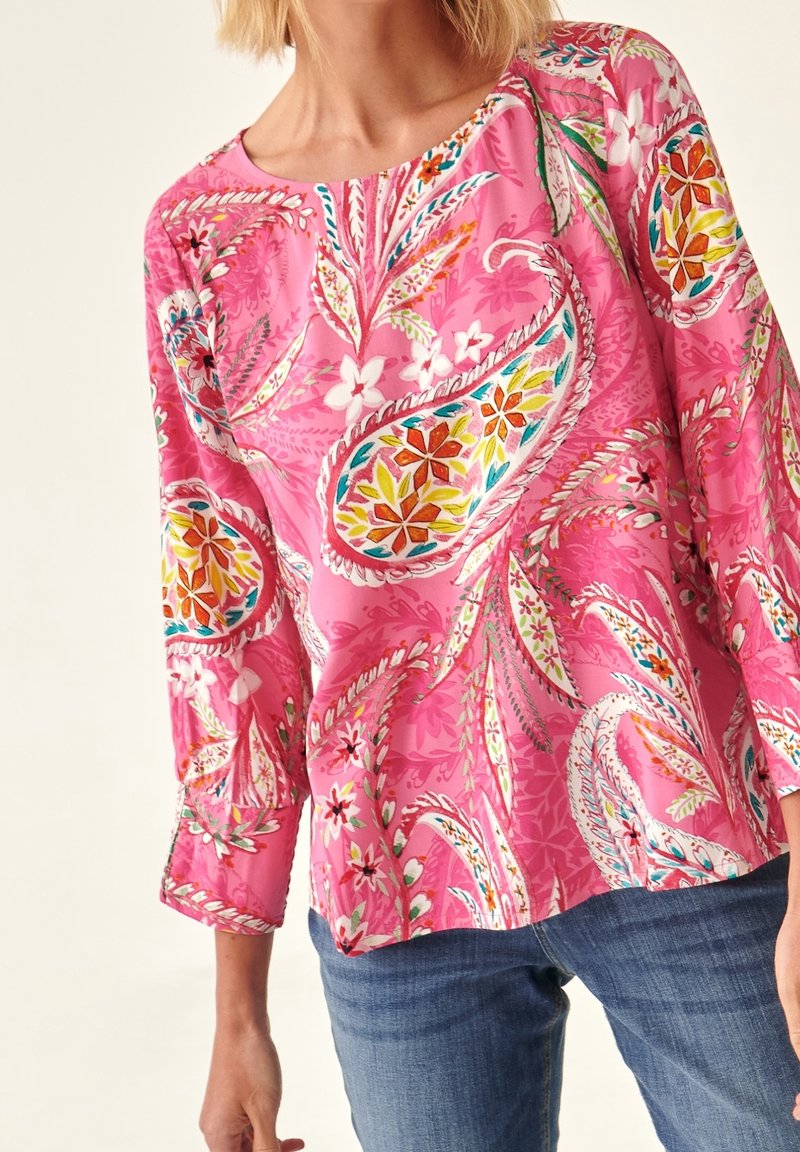 Femme portant un chemisier à manches longues rose vif avec des motifs floraux et cachemire colorés, associé à un jean bleu.