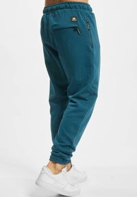 Tealfargede joggebukser laget av mykt stoff med elastisk midje, glidelåslommer og innsnevrede ermer, kombinert med hvite joggesko.