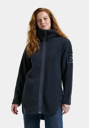 Jeune femme aux longs cheveux roux portant une veste polaire bleu marine foncé avec une fermeture éclair devant, se tenant devant un fond uni.