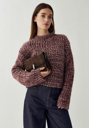 Giovane donna con capelli castani lunghi indossa un maglione spesso a maglia bordeaux e bianco, jeans scuri e tiene una pochette in camoscio marrone.