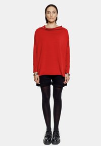 Czerwona bluzka z długim rękawem i drapowanym dekoltem, w połączeniu z czarnymi krótkimi spodniami z mankieciami, czarnymi rajstopami i czarnymi sznurowanymi butami. Minimalistyczne akcesoria.