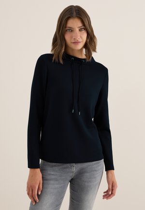 Kobieta o brązowych włosach do ramion, ubrana w czarną bluzę z długim rękawem i jasnoszare jeansy, stojąca na jednolitym tle.