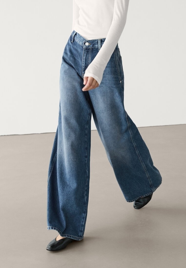 Jeans denim a gamba larga in blu medio con sbiaditure. Vita alta con chiusura a bottone e tasche laterali. Indossati con un top fitted a maniche lunghe bianco.