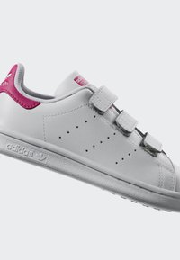 Baskets en cuir blanc avec trois bandes Velcro, accents roses sur le talon et la langue, détail perforé sur le côté et semelle en caoutchouc texturée.