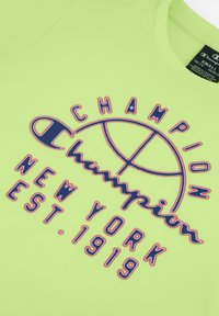 Camiseta gráfica verde con el logo de "Champion" y "Nueva York EST. 1919" en azul y rojo. Hecha de tela texturizada con un escote tipo crew.