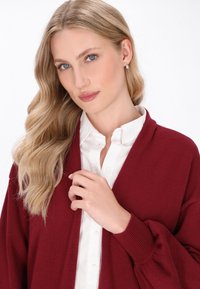 Cardigan bordeaux con scollo a V, polsini a costine e una texture morbida, indossato sopra una camicia bianca a maniche lunghe. Il modello tiene delicatamente il tessuto.