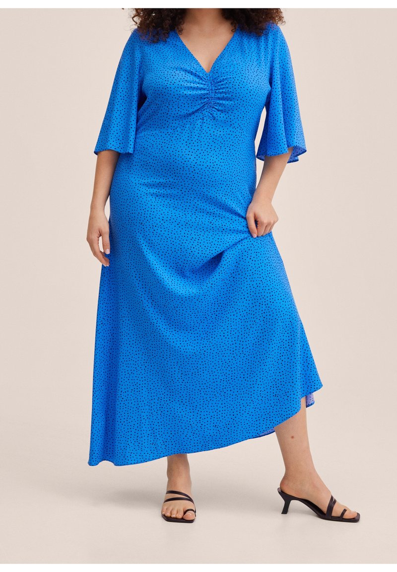 Robe longue bleue avec manches cloche courtes, décolleté en V, détail froncé au niveau de la poitrine, et pois noirs. Portée avec des sandales à talons noires.