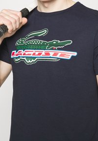 Mörkblå t-shirt i bomull med en grön krokodilgrafik och "LACOSTE"-logotyp i röd och vit över bröstet. Kort ärm.