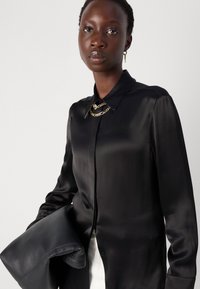 Camicia nera di raso a maniche lunghe con colletto a punta, caratterizzata da un dettaglio in catena dorata all'collo, tenuta con una borsa in pelle scura.