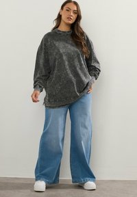 Felpa over-size grigio scuro con lavaggio acido, colletto alto e maniche con polsini, abbinata a jeans a gamba larga blu chiaro e sneakers bianche.