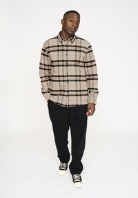 Beige kariertes Hemd mit schwarzen Streifen, Knopfverschluss vorne und langen Ärmeln. Kombiniert mit schwarzen Hosen und schwarz-weißen Turnschuhen.