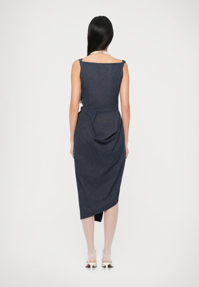 Mujer con cabello largo y oscuro vestida con un vestido asimétrico sin mangas de color azul oscuro, tacones blancos y un collar de perlas, de espaldas sobre un fondo blanco.