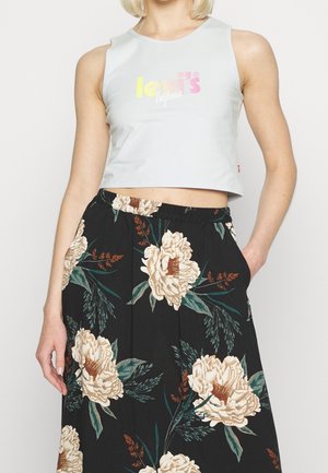 Mujer con top corto blanco con logo en tonos pastel y falda floral negra de talle alto con flores beige y hojas verdes.