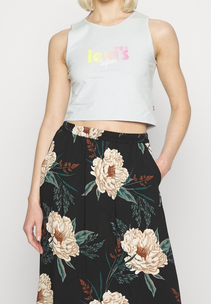 Femme portant un top court blanc avec un logo pastel et une jupe fleurie noire taille haute avec des fleurs beiges et des feuilles vertes.
