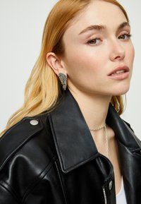 Pendientes plateados con forma de ala y detalles texturizados, llevados por una persona con chaqueta de cuero negra. Collar sencillo de cadena visible.