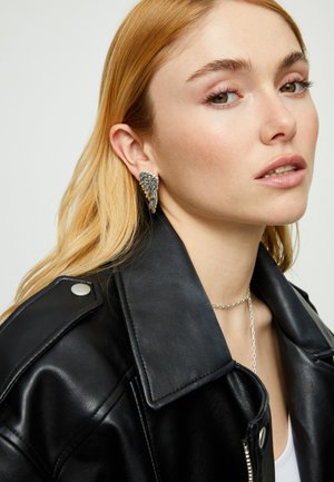 Pendientes plateados con forma de ala y detalles texturizados, llevados por una persona con chaqueta de cuero negra. Collar sencillo de cadena visible.
