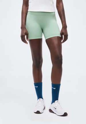 Person iført lysegrønne atletikshorts, blå Nike mellemlange sokker, hvide Nike sneakers og en hvid top mod en ensfarvet lys baggrund.