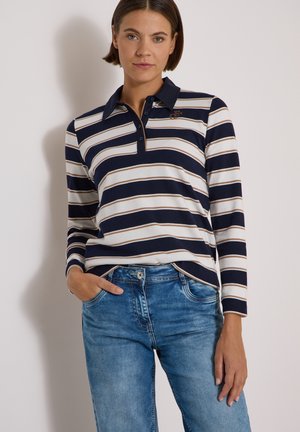 Frau mit kurzen braunen Haaren, die ein navyblaues, weißes und beiges horizontal gestreiftes Langarm-Poloshirt und blaue Jeans trägt, steht mit einer Hand in der Tasche.