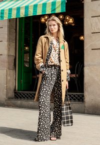 Béžový trenčkot na čiernej a bielej blúzke s bodkami a zodpovedajúcich širokých nohaviciach, držiaci károvanú kabelku. Zelené dvere v pozadí.