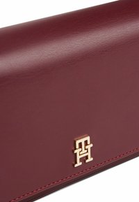 Burgunderfarbige Lederbörse mit strukturierter Oberfläche, versehen mit einem goldenen Logo-Detail und kontrastierenden roten Nähten entlang der Kante.