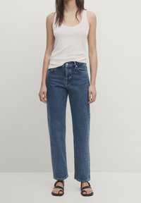 Vit ärmlös linne som kombineras med högmidjade blå denimjeans och svarta platta sandaler. Texturen är slät med en avslappnad passform.