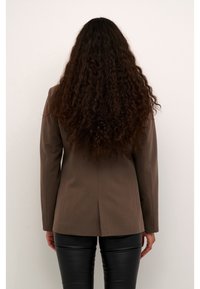 Blazer marrón ajustado hecho de una tela suave, con una abertura única en la parte trasera, combinado con pantalones de cuero negro brillante.