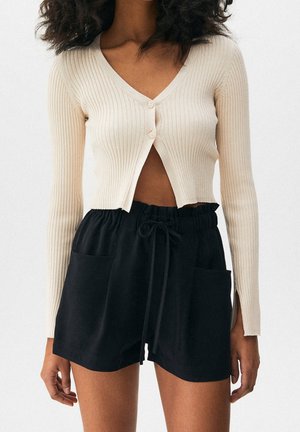 Kvinde iført en beige ribstrikket cropped cardigan med lange ærmer og to knapper samt sorte shorts med snøre og forlommer.