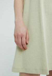 Ljusgrön klänning i mjuk bomull, med avslappnad passform, korta ärmar och rundad fåll, med en hand som vilar mjukt mot tyget.