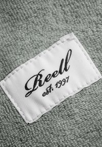 Green terry cloth fabric features a white label stitched on with black cursive text reading "Reell est. 1997." Soft texture and neat stitching visible.