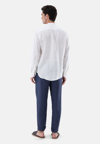 Weißes Leinenhemd mit langen Ärmeln und abgerundetem Saum, kombiniert mit blauen Leinenhosen und dunklen Sandalen. Die Rückansicht zeigt eine lässige Passform.