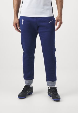Mann trägt dunkelblaue Sporthose mit Tottenham Hotspur und Nike Logos, schwarze Fußballschuhe, weiße Socken und ein weißes Hemd, das am Bund sichtbar ist.