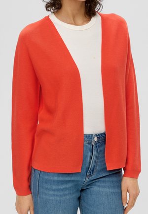 Femme portant un cardigan orange vif ouvert sur un top blanc et un jean bleu sur un fond clair uni.