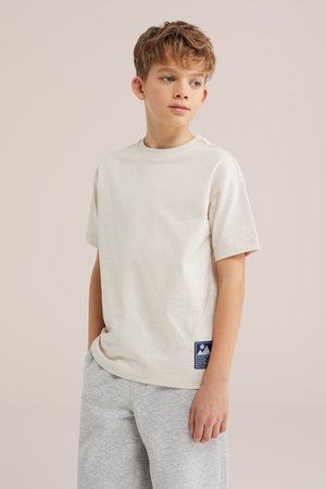 Vaaleahiuksinen poika, joka käyttää yksinkertaista off-white -väristä t-paitaa, jossa on sininen paikka, ja vaaleanharmaita shortseja, katsoo ajatuksissaan sivulle.