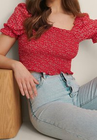 Rotes Blumen-Crop-Top mit kurzen Puffärmeln, das über eine geruckte Textur verfügt. Kombiniert mit hochgeschnittenen hellblauen Jeans.