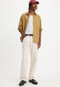 Lichtbruine overhemd met knopen over een wit tanktop, crème broek en zwarte schoenen. Accessoires zijn een kastanjebruine pet en een gestreepte riem.