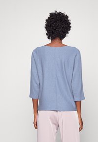 Ljusblå tröja med fyrkantig halsringning, trekvartslånga ärmar och textilstruktur. Har en diskret söm längs mitten av ryggen.