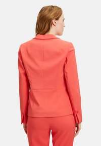 Korálový blazer s klopami a strukturovanými rameny. Hladká látka, přiléhavý design a knoflíkové detaily na manžetách. Zobrazeno vzadu.