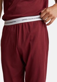 Osoba v burgundských kalhotách a tričku upravuje bílý elastický pás označený "United Colors of Benetton."
