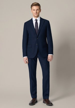 Mężczyzna stoi, ubrany w ciemnogranatowy garnitur, białą koszulę, ciemny krawat oraz brązowe skórzane eleganckie buty, na tle jednolitego beżowego tła.